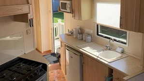 A compact kitchen with a gas stove, oven, refrigerator, and a sink with a window above it. - Beach Vibes - Silver Beach - Basic (Skegness)