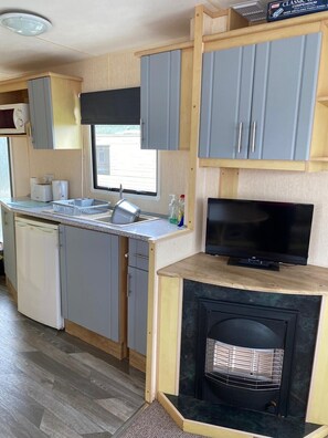 A compact kitchen with grey cabinets, a built-in dishwasher, a small sink, a window, a flat-screen TV, and a fireplace. - Beach Vibes - Silver Beach - Basic (Skegness)