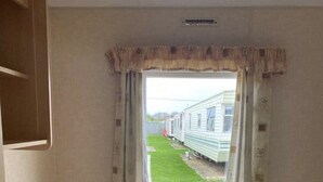 A small, single-room RV with two beds, a window with curtains, and a view of other RVs outside. - Beach Daze - Silver Beach - Standard (Skegness)