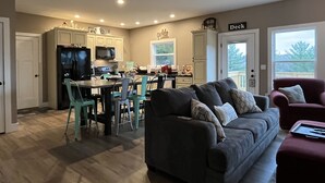 A spacious living room with a dark grey sofa and teal chairs, a kitchen with white cabinets and a black refrigerator, and a deck area with a sign reading "Deck". - FAMILY LAKE ESCAPE AWAITS. Walk to Boat Ramp. Sleeps 12, with plenty of parking (Bronston)