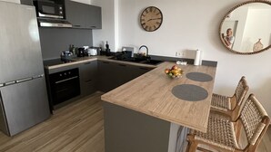 A modern kitchen with a central island, built-in appliances, and a dining area with a woven chair. - Bella Vista 2 bedroom - Orient Bay (Collectivité de Saint-Martin)