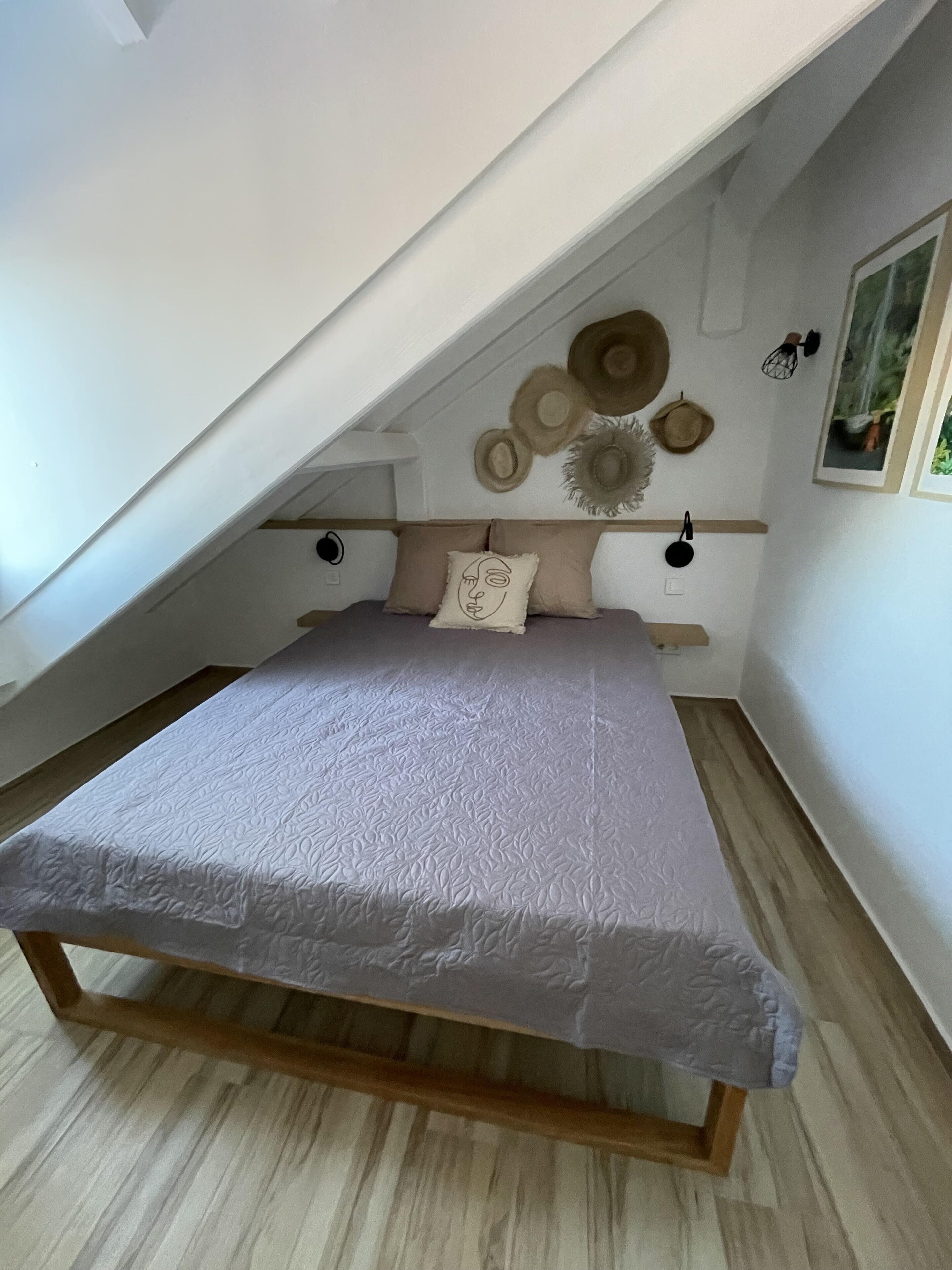 Una camera da letto con soffitto spiovente, un letto con copripiumino bianco testurizzato, due cuscini, una mensola a parete con cappelli e un quadro incorniciato.