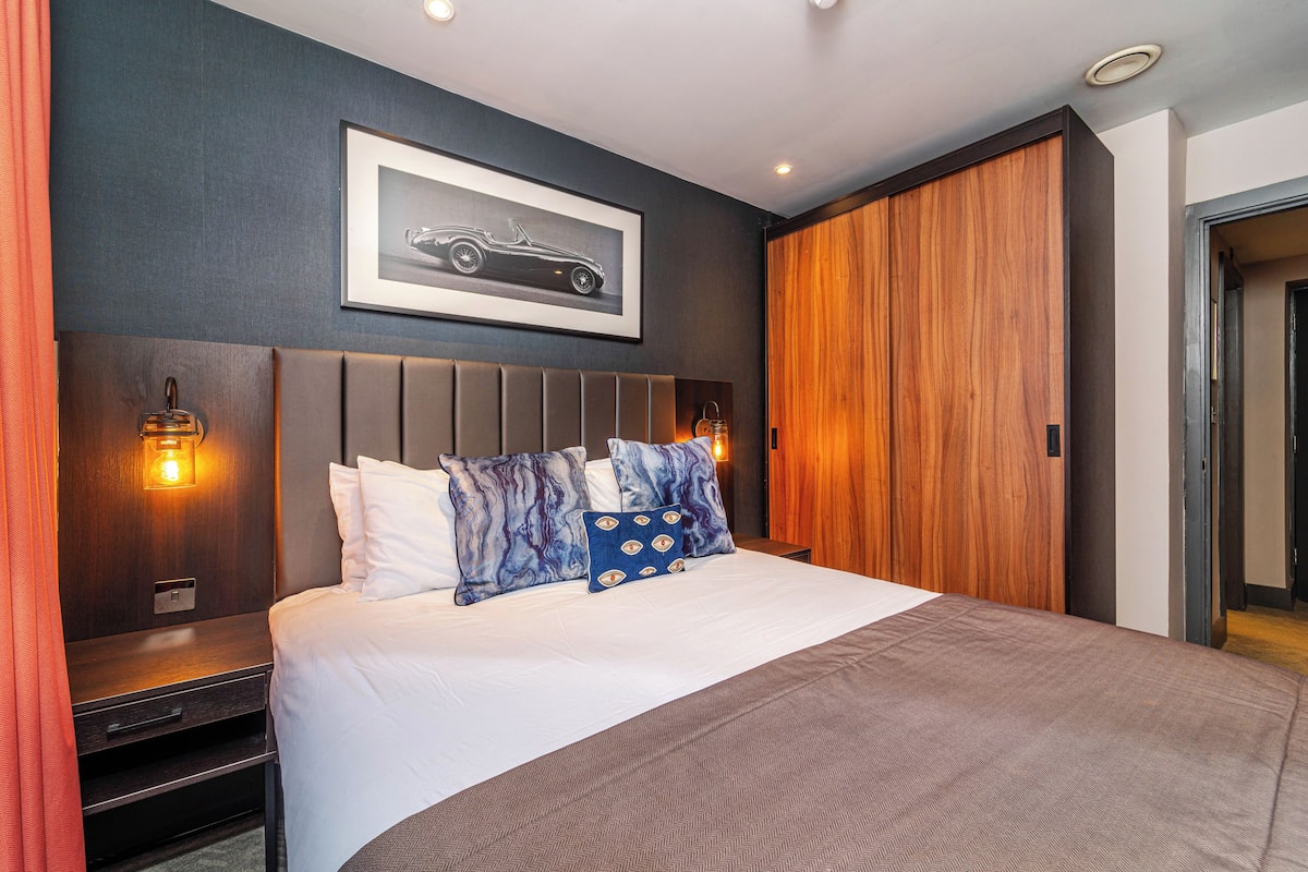 A modern hotel room with a large bed, a headboard with built-in lighting, a framed picture on the wall, and a wooden wardrobe.