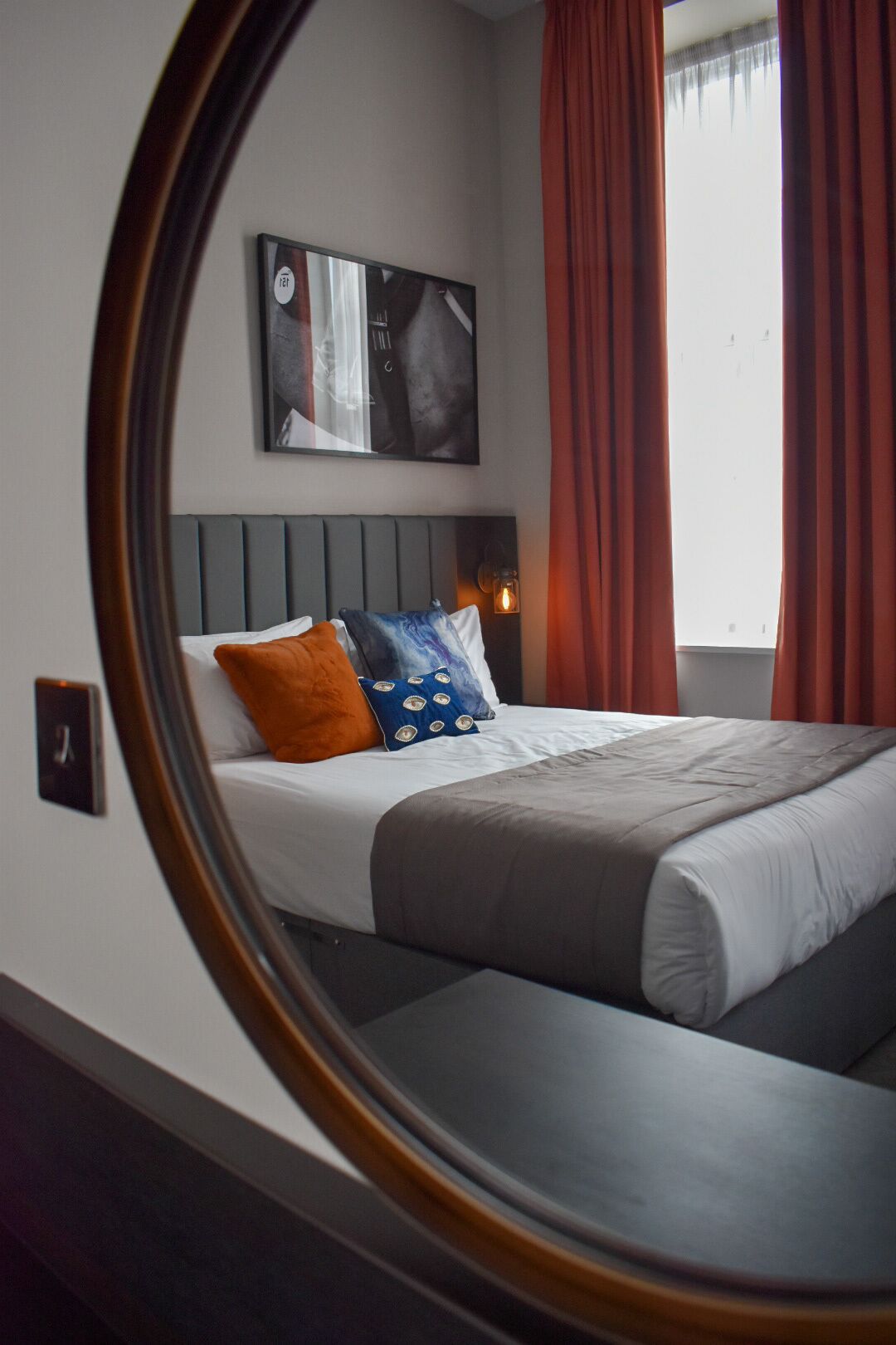 A bedroom with a bed, pillows, a headboard, a wall-mounted picture, and red curtains.