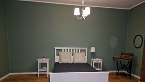 A bedroom with a wooden floor, a white bed with two pillows, a nightstand with a lamp, and a vintage sewing machine on a table. - Beautiful place in the city center (Kraków)