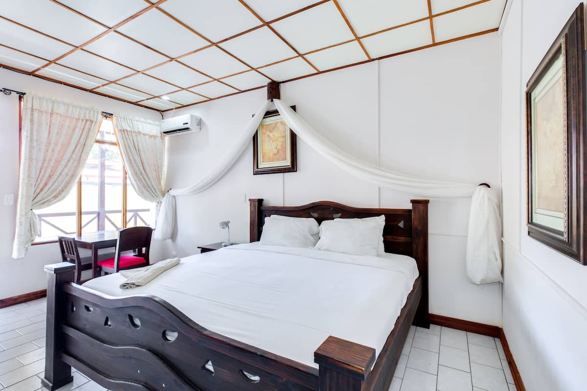 A neatly arranged bedroom with a wooden bed, a desk, and chairs, a window with curtains, and a framed picture on the wall.