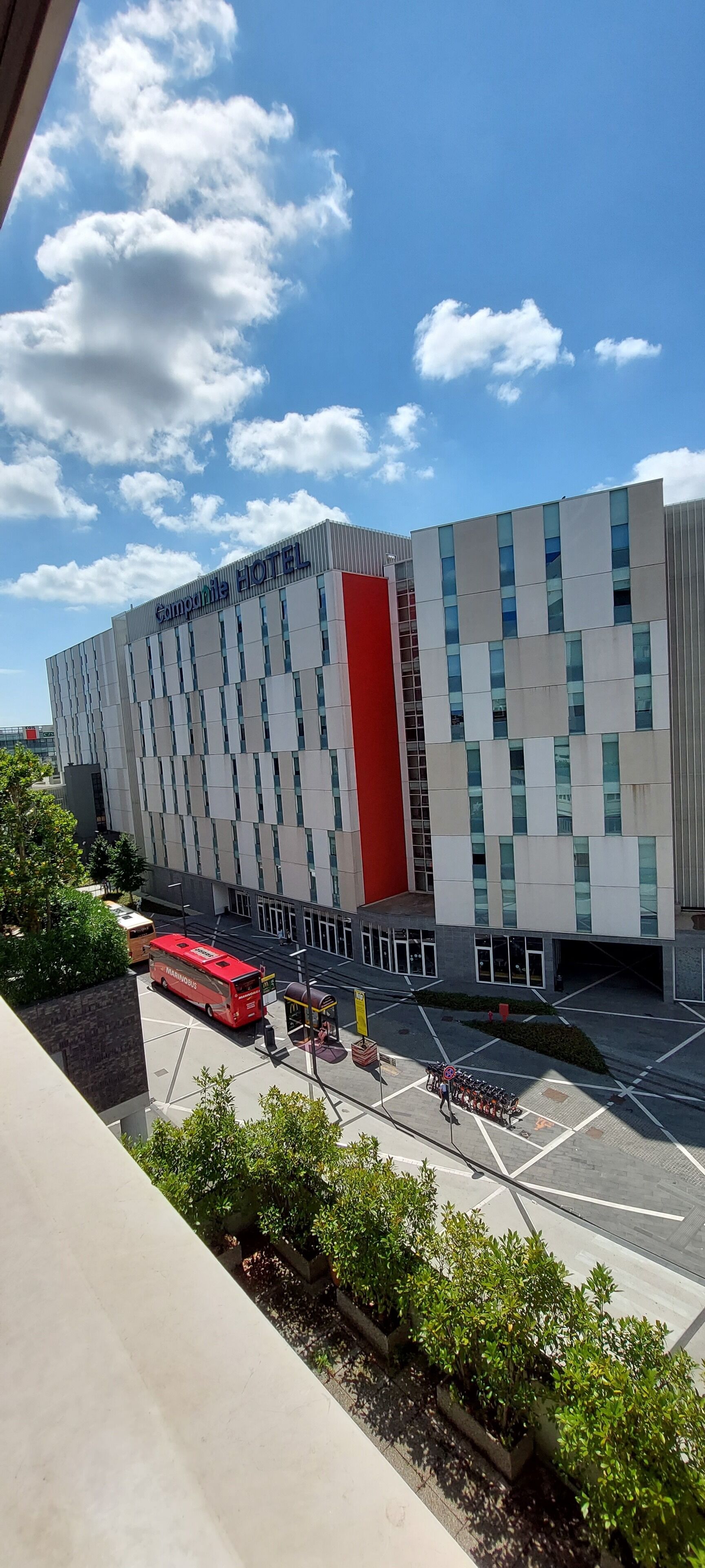 A modern hotel building with a prominent red vertical stripe, large windows, and a parking area with a red bus.