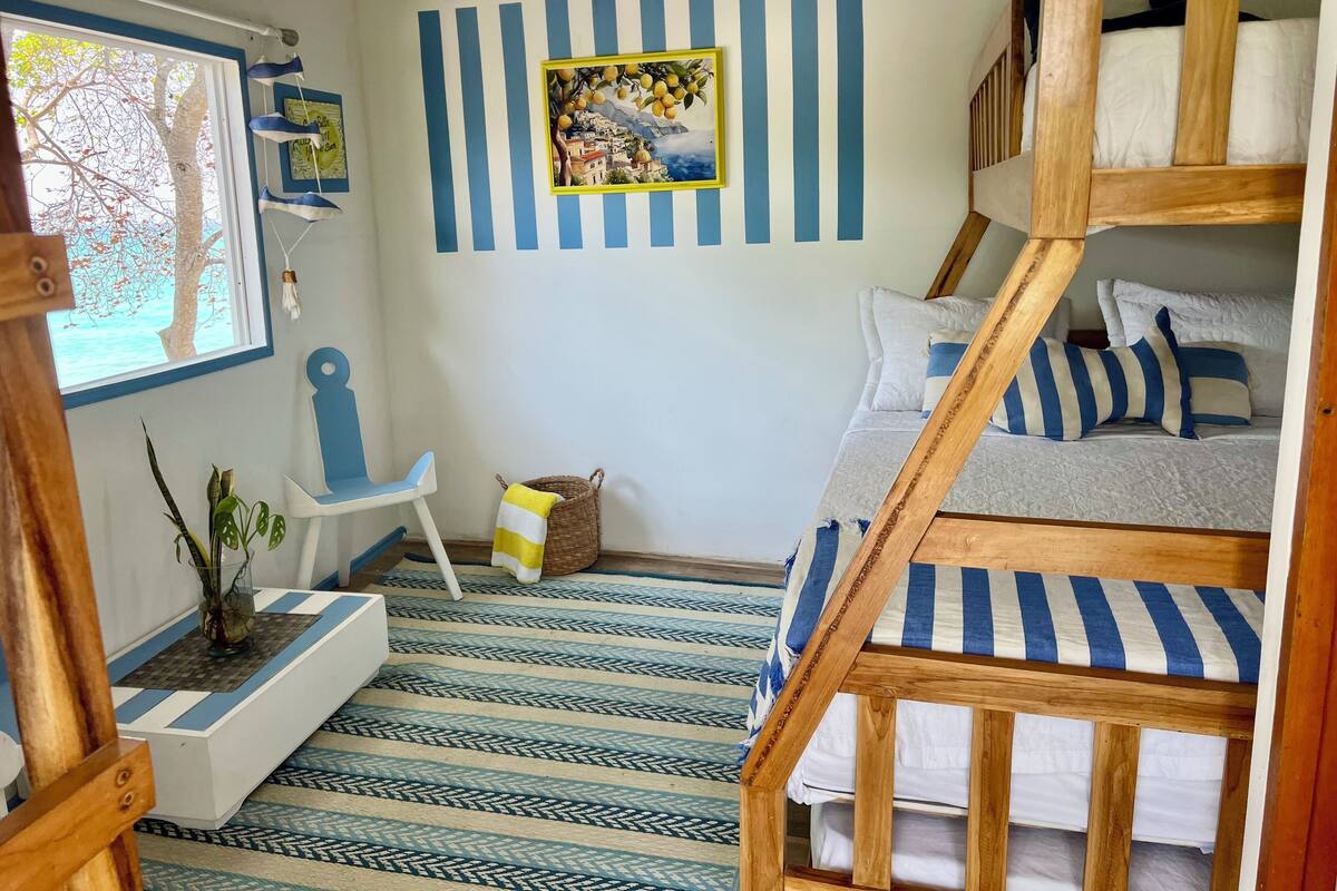 A room with a bunk bed, a small desk, a chair, a window with a view of trees, and a wall with a striped pattern and a framed picture.