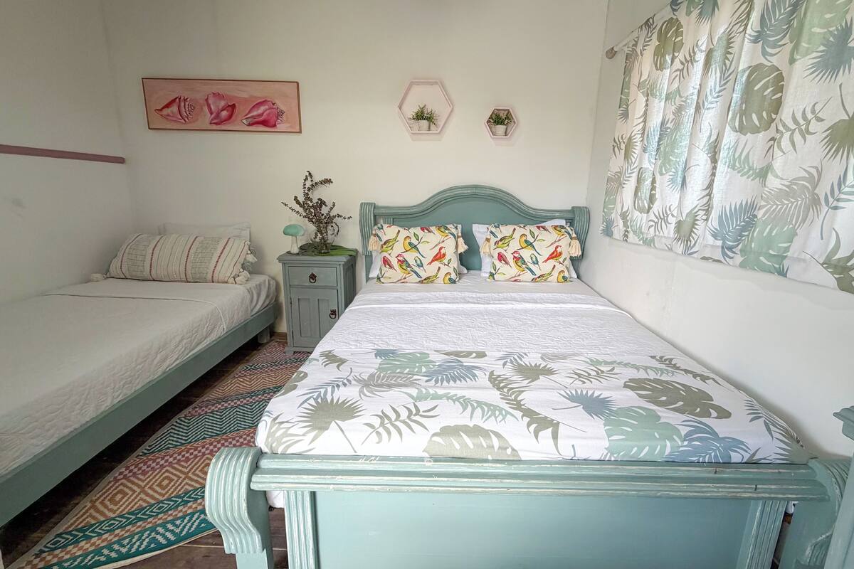 A bedroom with two beds, a nightstand, a dresser, a patterned rug, and a wall with a floral painting and two hexagonal shelves with plants.