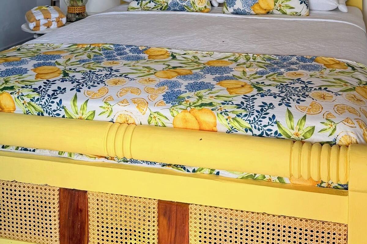 A bed with a yellow headboard and floral bedding, a potted plant, and three framed artworks on the wall.