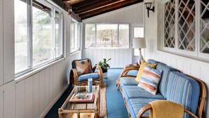 Ein gemütliches Zimmer mit Holzbalkendecke, großen Fenstern, Korbmöbeln und einem blauen Teppich.