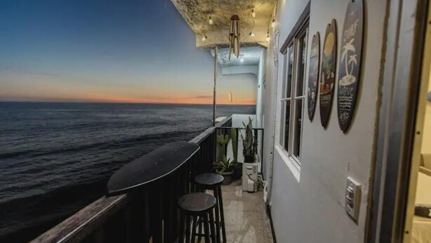 Un balcón con vista al océano al atardecer, que incluye una zona de bar con taburetes y una lámpara colgante.