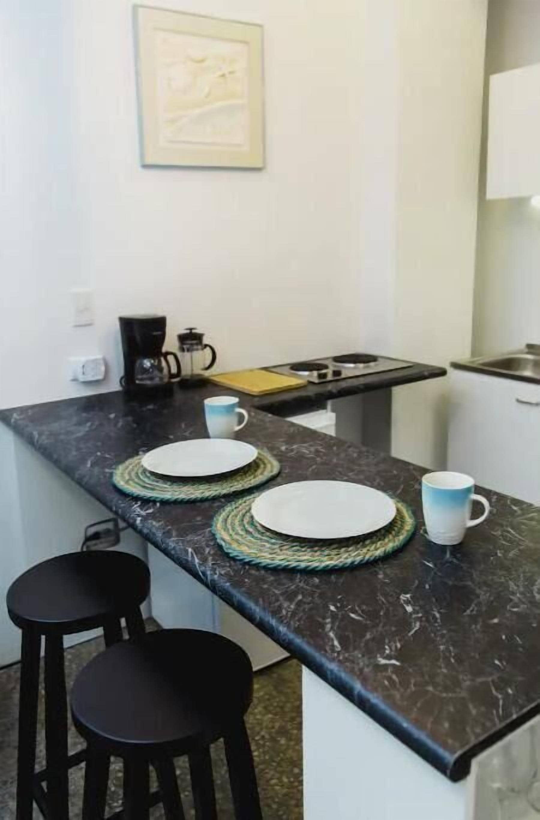 A small kitchen with a marble countertop, two stools, a coffee maker, and a framed picture on the wall.