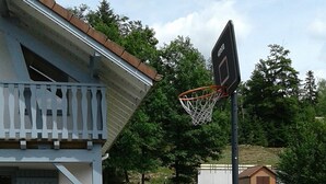 Zona residencial con un aro de baloncesto, una casa con balcón y un cobertizo pequeño al fondo.