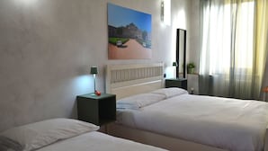 Une chambre d’hôtel avec deux lits simples, une table de chevet avec une lampe, un tableau au mur et une fenêtre avec des rideaux.