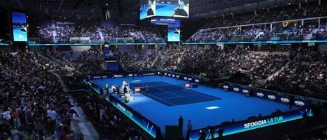 A large indoor sports arena with multiple screens, a tennis court, and a crowd of spectators.