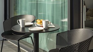 Una mesa redonda con dos tazas blancas, un plato con cruasanes y un periódico. La mesa está colocada en un balcón con barandilla de cristal y vistas al exterior.