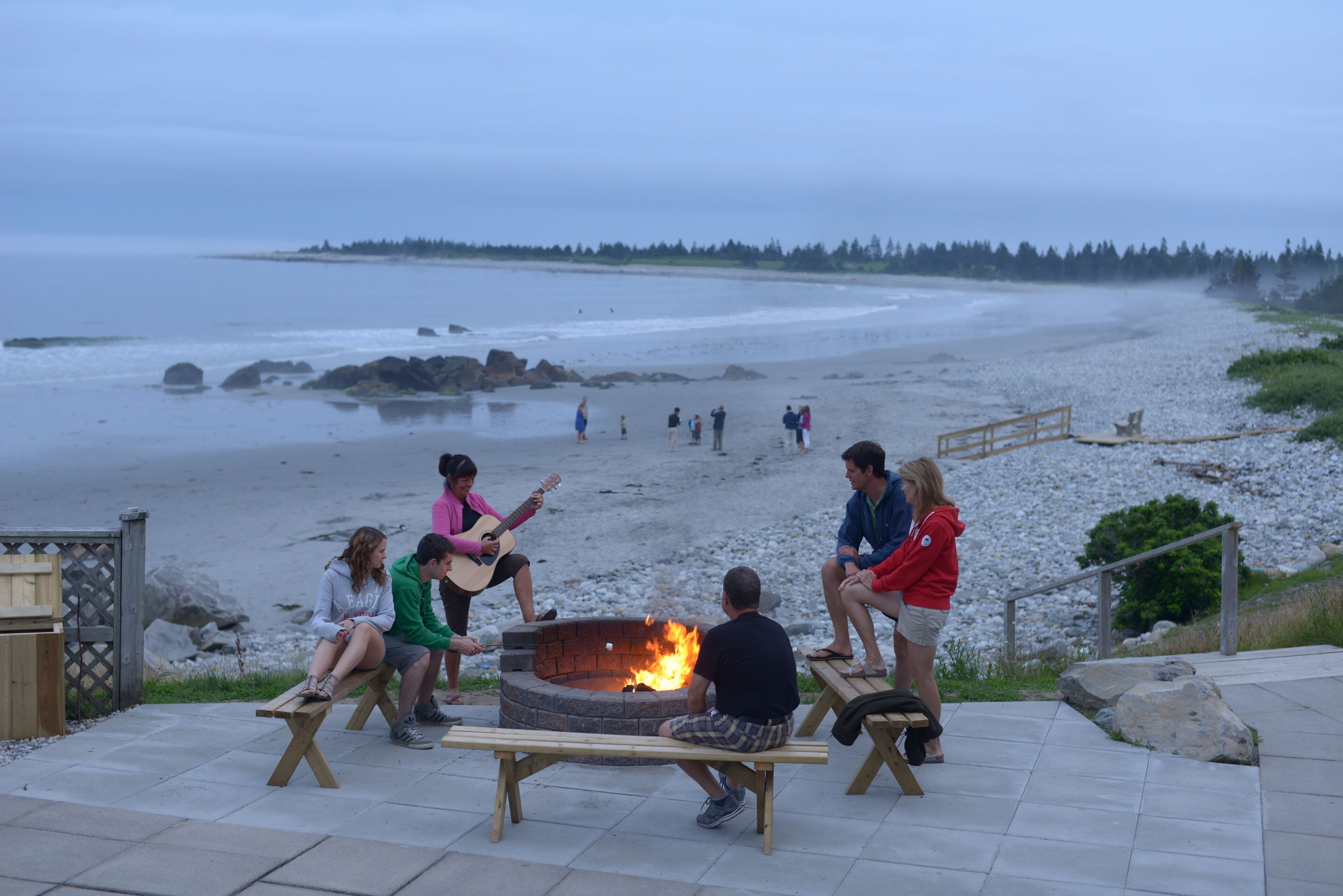 Un groupe de personnes est assis autour d’un foyer sur une plage, avec une vue sur l’océan et une forêt en arrière-plan.