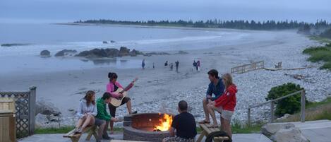 Un groupe de personnes est assis autour d’un foyer sur une plage, avec une vue sur l’océan et une forêt en arrière-plan.