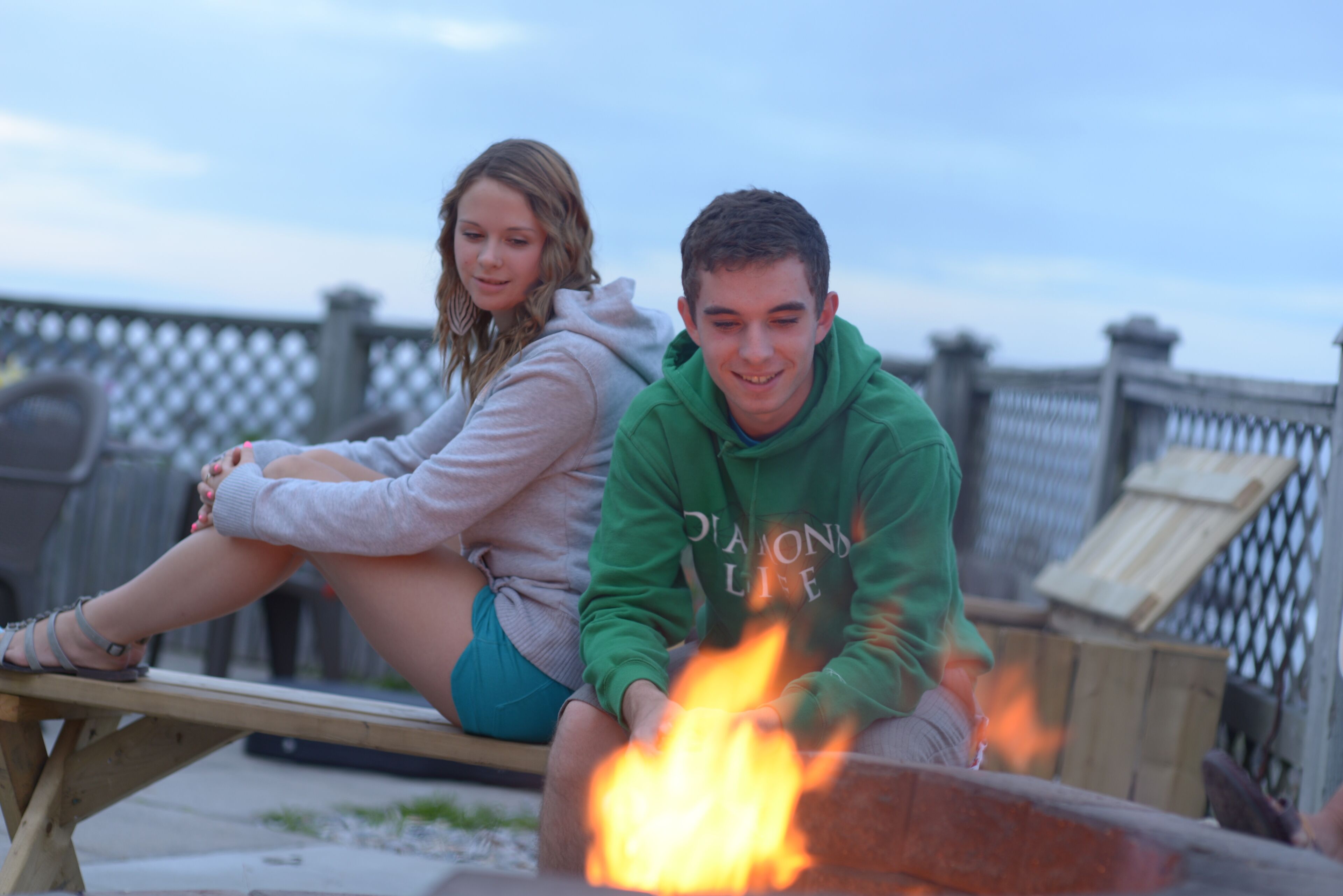 Two people sitting by a fire pit on a wooden deck with a metal fence in the background.