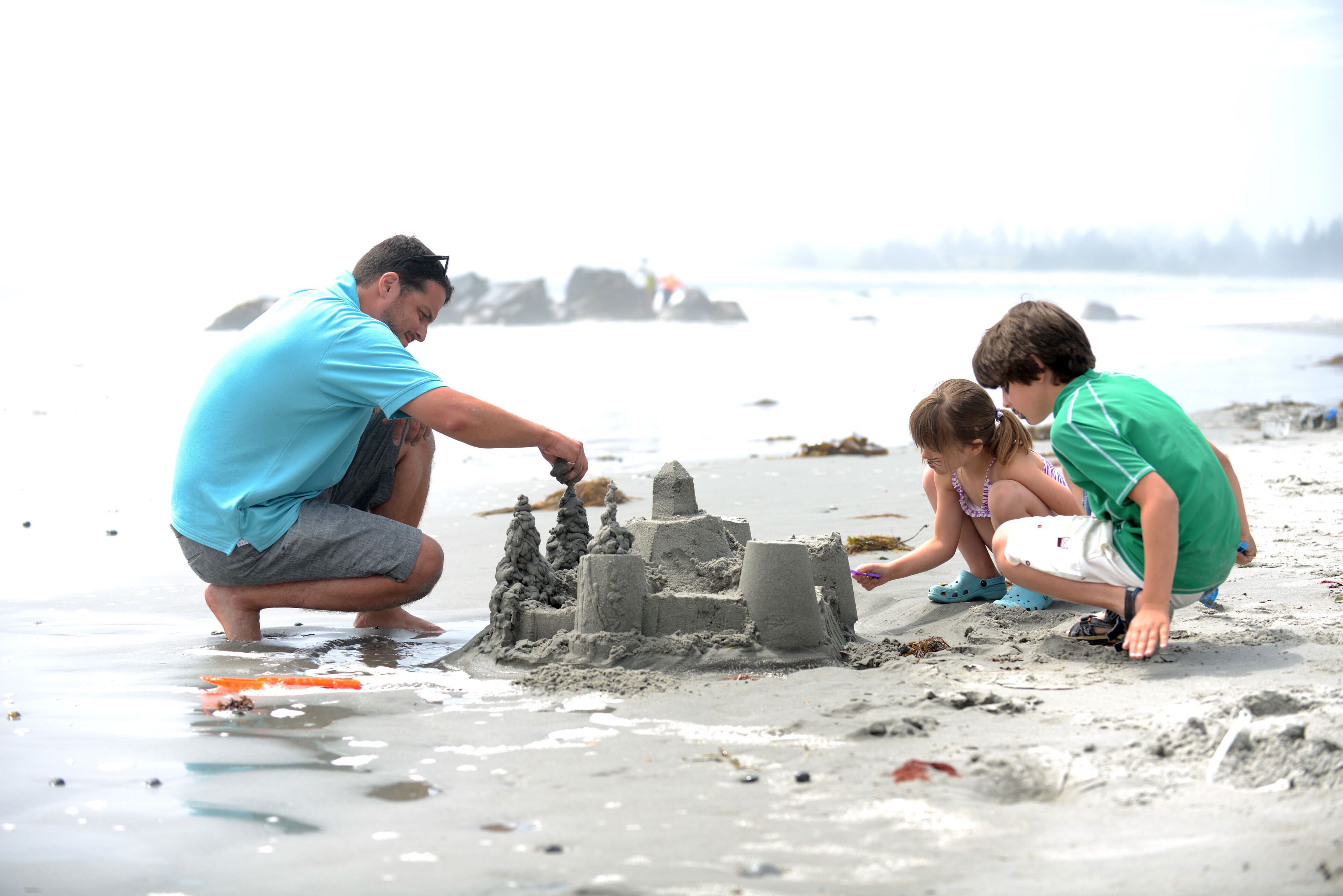Una playa arenosa con un castillo de arena que tres personas están construyendo.