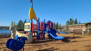 A playground with a slide and climbing structures, located near a body of water and a building.