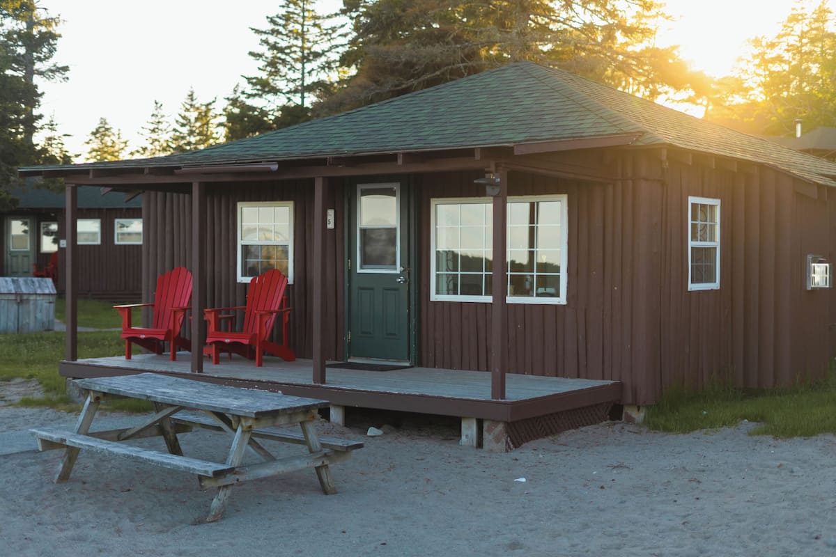 Una piccola casetta di legno con un portico, due sedie Adirondack rosse e un tavolo da picnic.