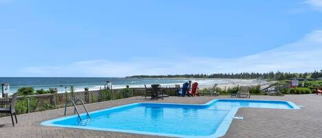 Une piscine rectangulaire aux bords bleus, entourée d’une terrasse en briques. On y trouve des chaises longues et une table près de la piscine, et une vue sur l’océan et la plage en arrière-plan.