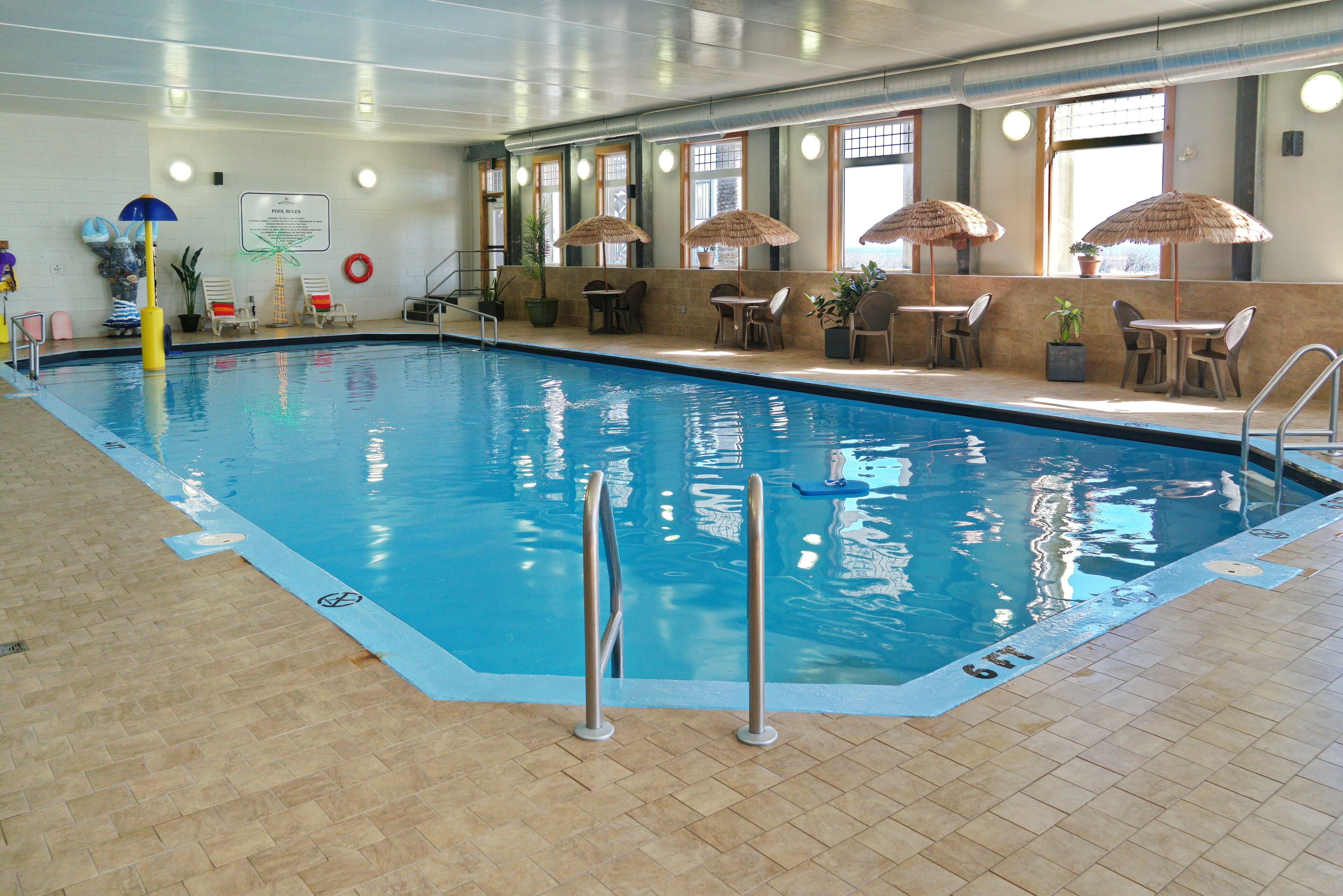 An indoor swimming pool with clear water, surrounded by lounge chairs and umbrellas, and a lifebuoy on the wall.