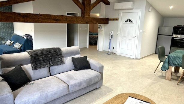 A modern living room with a grey sofa, wooden beams, a dining area with a table and chairs, and a kitchen in the background. - Le Cosy de Pauline et Flo (Cessenon-sur-Orb)