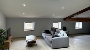A modern living room with a grey sofa, a wooden coffee table, and a TV on a wooden stand. There are two windows providing natural light, and the ceiling has exposed wooden beams. - Le Cosy de Pauline et Flo (Cessenon-sur-Orb)