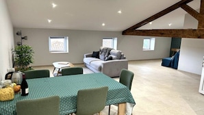A spacious room with a high ceiling, a dining table with chairs, a sofa, and a small table with a plant and a bottle of wine. - Le Cosy de Pauline et Flo (Cessenon-sur-Orb)