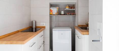 A compact kitchen with white cabinets, a wooden countertop, a white refrigerator, and a shelf with kitchen items.