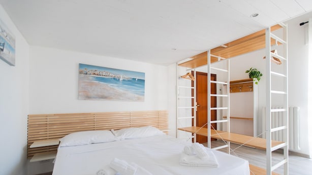A modern hotel room with a large bed, wooden headboard, a painting of a coastal scene, and a built-in wardrobe with a plant.
