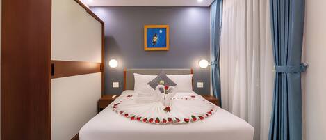 A neatly made bed with a swan design and rose petals, a wooden wardrobe, a nightstand with a lamp, a framed picture on the wall, and a window with blue curtains.