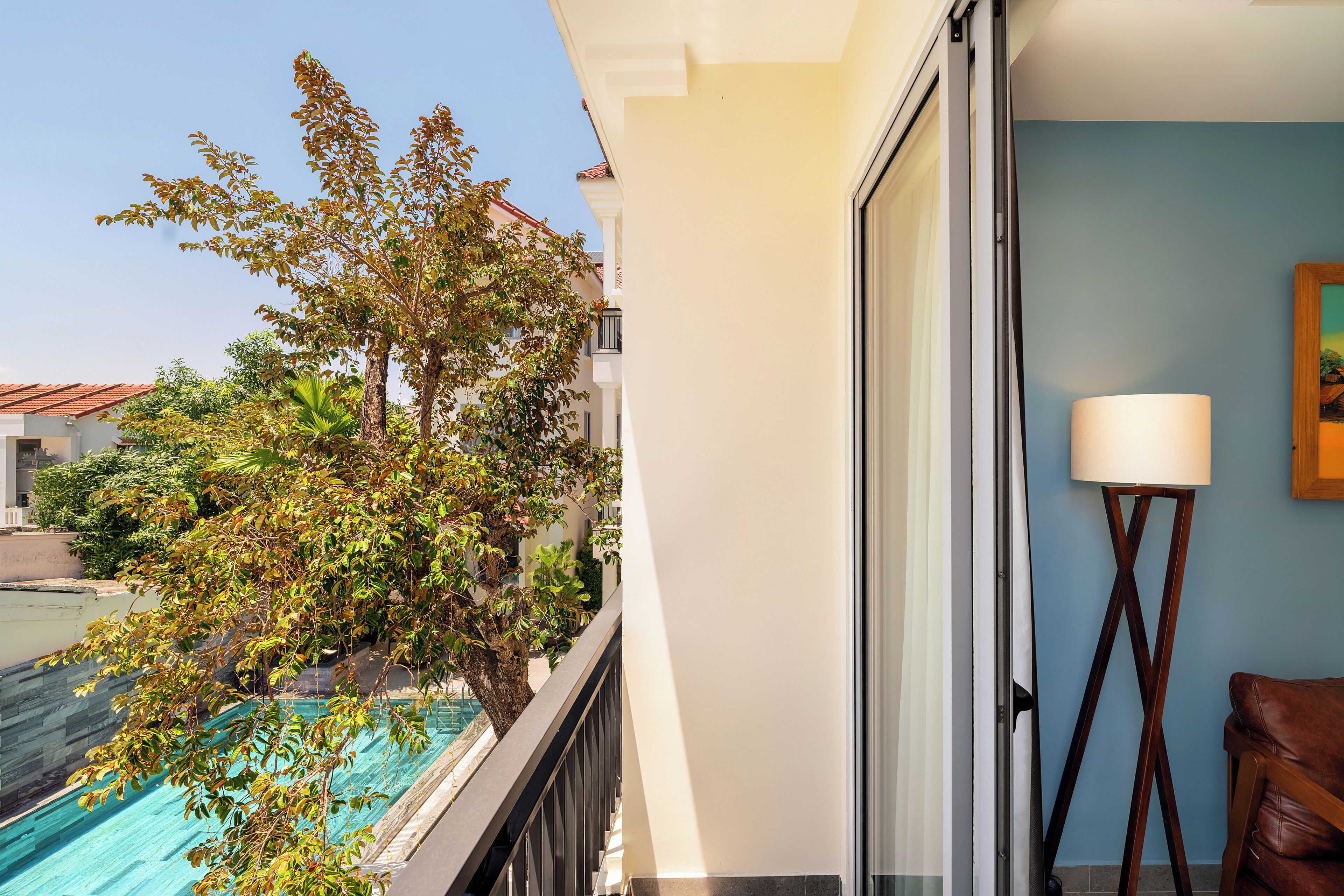 A balcony with a view of a swimming pool and trees, a lamp, and a chair.