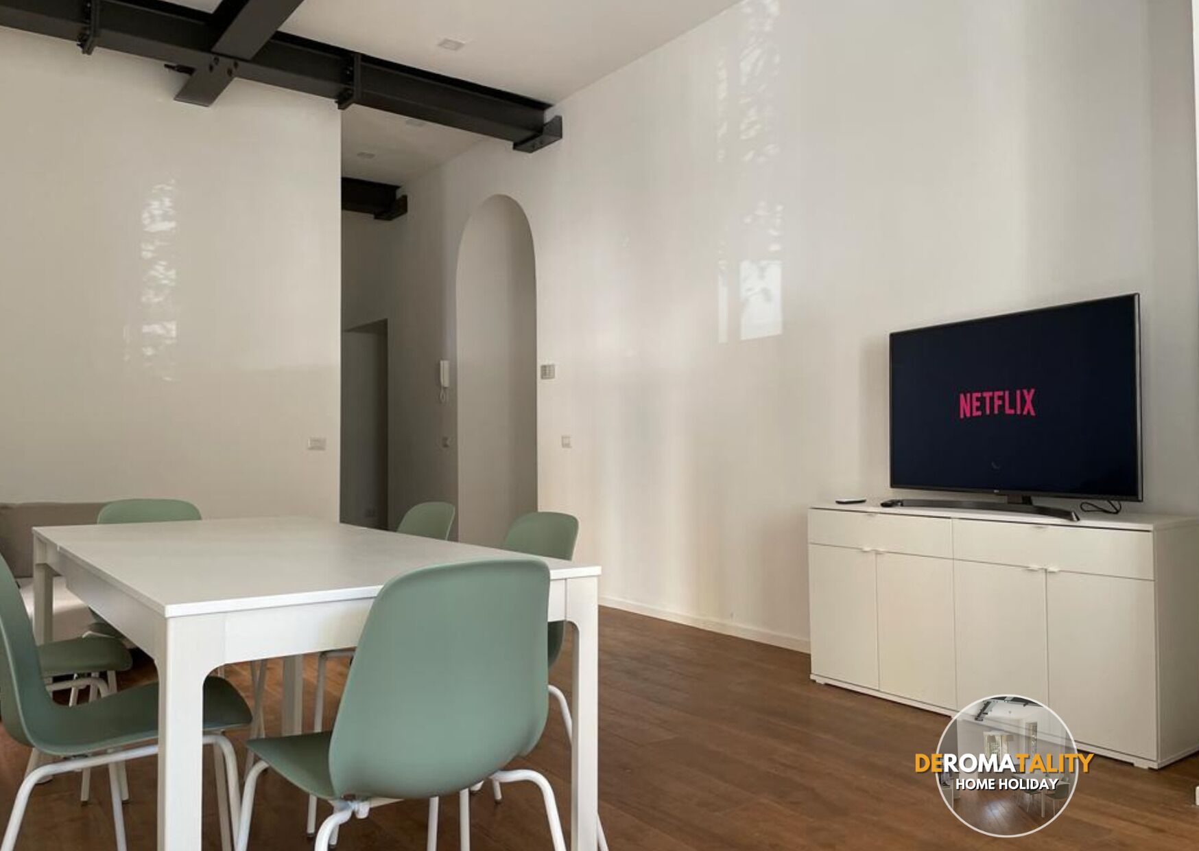Una sala moderna con una mesa blanca y sillas verdes, un televisor de pantalla plana mostrando 'NETFLIX' y un mueble blanco debajo.