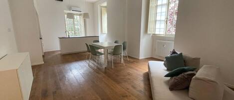 A spacious living area with a wooden floor, a dining table with chairs, a sofa with cushions, and large windows providing natural light.