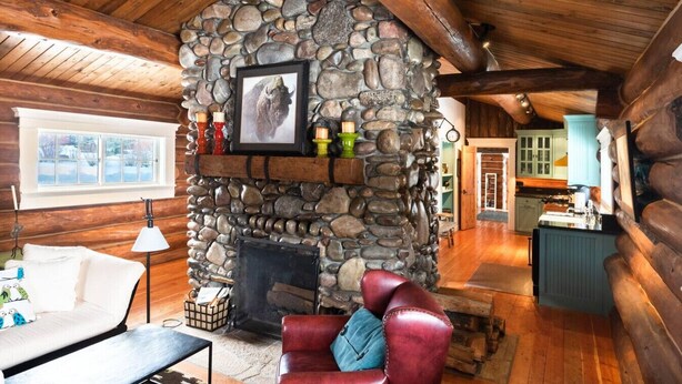 A cozy cabin interior with a stone fireplace, wooden beams, a plush red leather sofa, and a view of the snowy outdoors through a window.