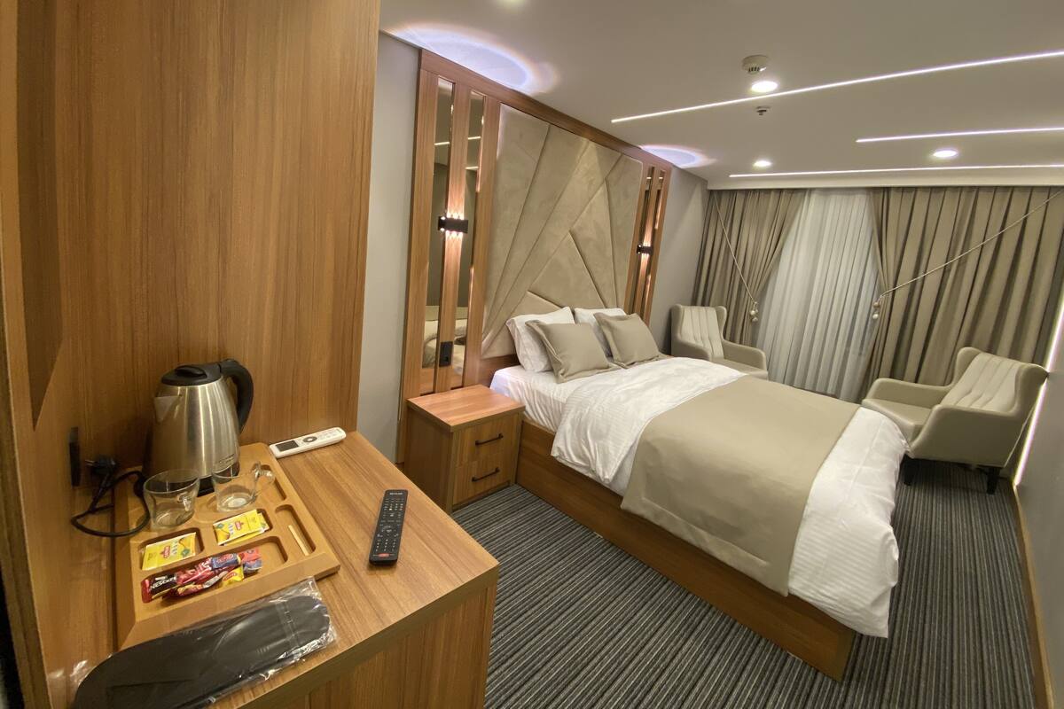 A modern hotel room with a wooden desk, a bed with a beige headboard, a bedside table with a lamp, and a chair.
