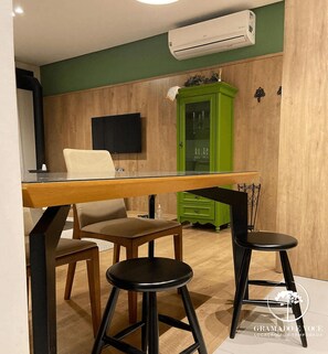 A dining area with a wooden table, two black bar stools, a green cabinet, and a mounted TV. - Apartamento Novo Loft One Centro Gramado (Gramado)
