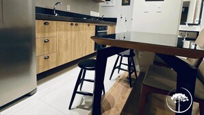 A modern kitchen with wooden cabinets, a stainless steel refrigerator, and a dining area with stools. - Apartamento Novo Loft One Centro Gramado (Gramado)