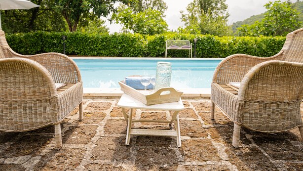 A wicker chair and table set by a pool with a glass vase and a tray of items.