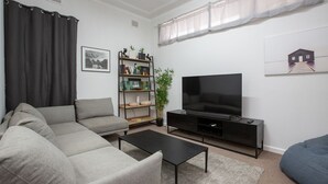 Moderni olohuone, jossa on harmaa sohva, musta sohvapöytä, mustalla jalustalla oleva televisio, kasveilla koristeltu kirjahylly ja seinällä oleva talokuva.