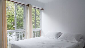 Un dormitorio con un ventanal amplio, una cama con ropa de cama blanca y vistas a árboles.