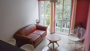 Un salón con un sofá de cuero rojo, una mesa redonda de madera y una silla blanca de alambre. Cuenta con un ventanal con cortinas, una lámpara colgante y una planta en maceta en el alféizar.