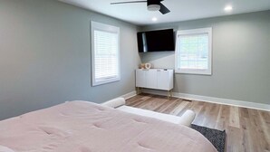 Une chambre à coucher avec un lit, un ventilateur de plafond, un téléviseur fixé au mur et une fenêtre avec des stores.