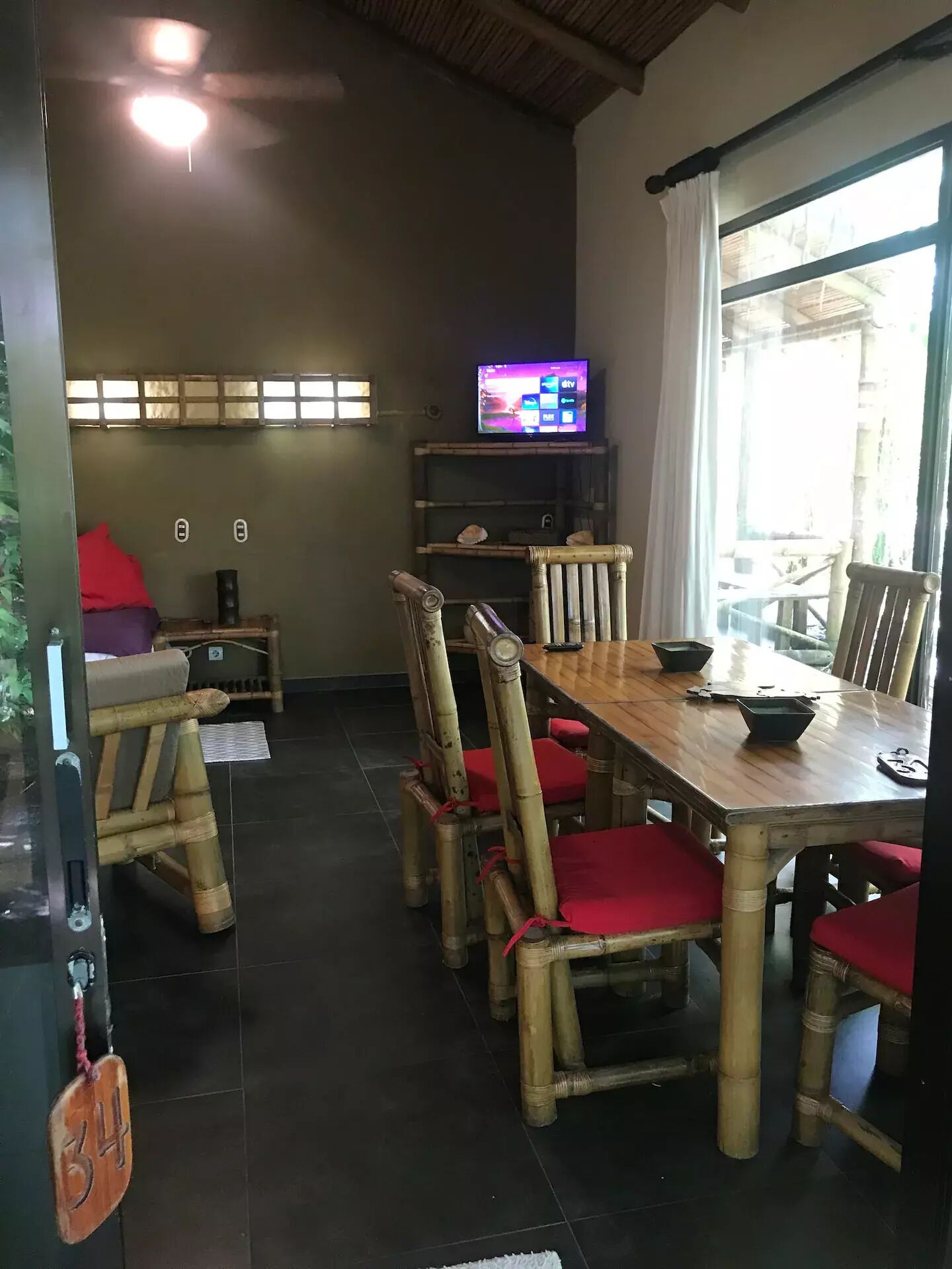 Un coin repas avec des chaises en bambou et une table en bois, un téléviseur posé sur une étagère et une fenêtre avec des rideaux.