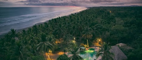 A resort with a pool surrounded by palm trees, overlooking a beach and ocean at sunset.