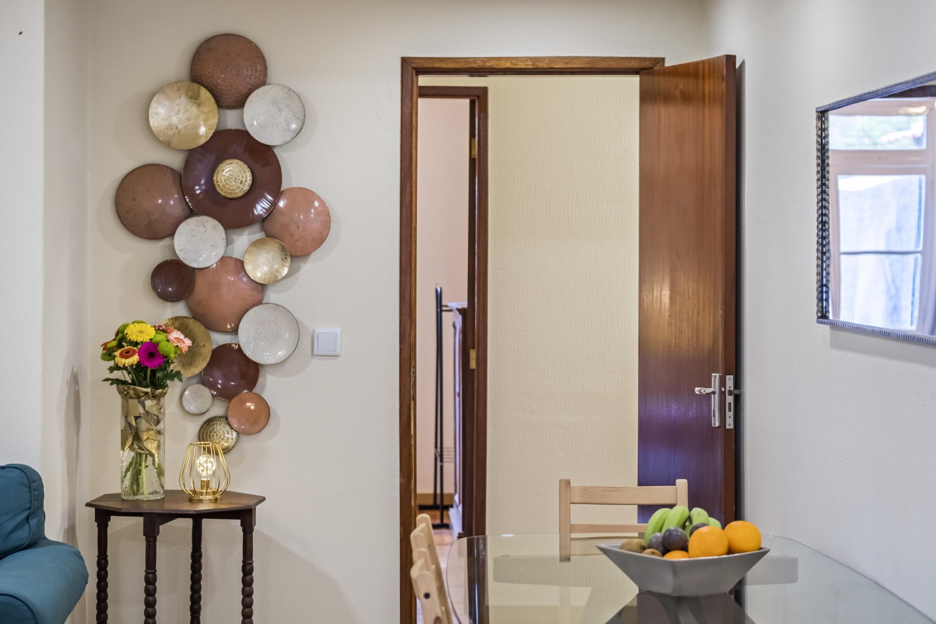 A room with a wooden door, a vase with flowers, a bowl of fruit, and a wall-mounted decorative plate arrangement.